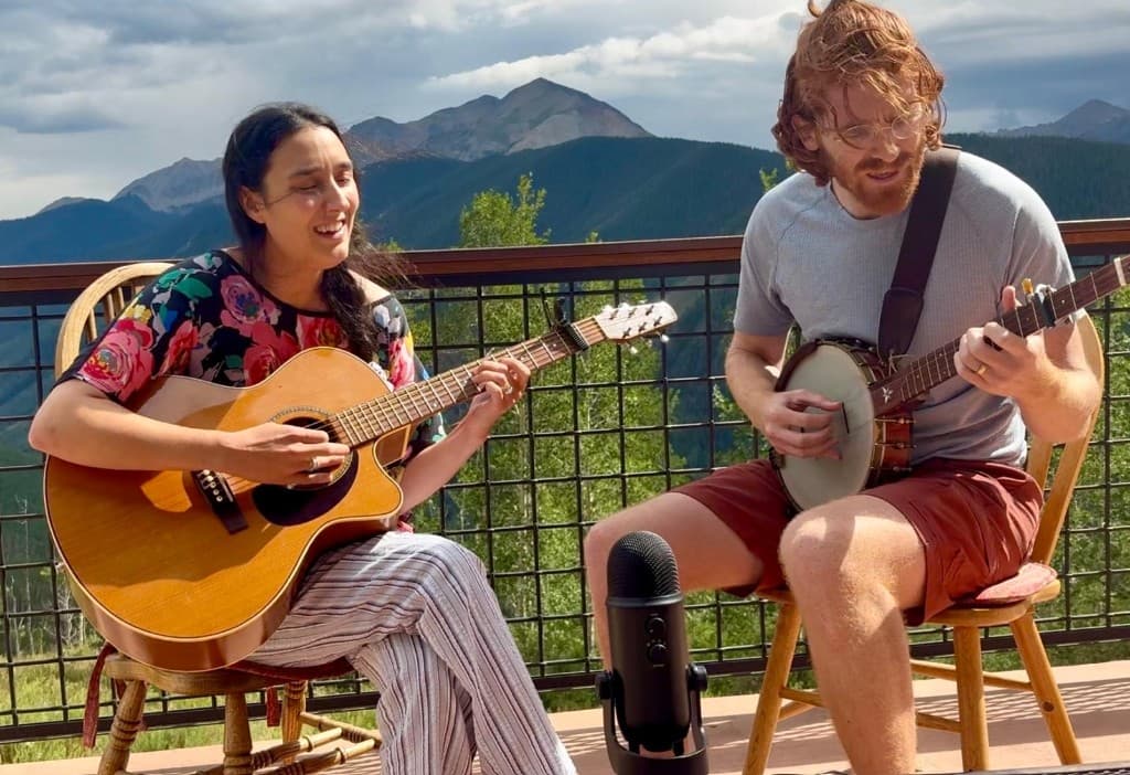Sarah and Ben playing guitar and banjo on a balcony with mountains behind